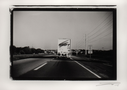 black and white image of a freight truck from behind traveling down a two lane highway. Deep shadows to the left.