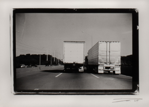 Black and white image of two side-by-side freight trucks as viewed from behind traveling down a highway.