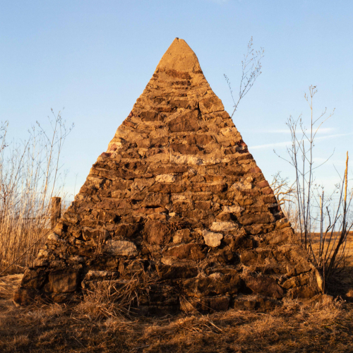 A large image of a stone and mortar pyramid in a leafless field with a bright blue sky