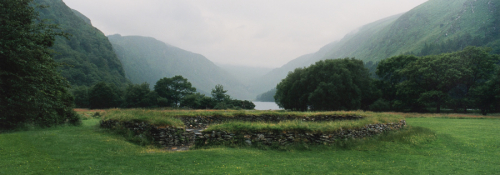  panoramic landscape image of a ring of stones covered in grass with a lake and misty mountains in the distance.
