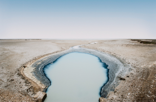 Image of a cloudy, gray pool of water (lower center) surrounded by a barren landscape below a cloudless sky.