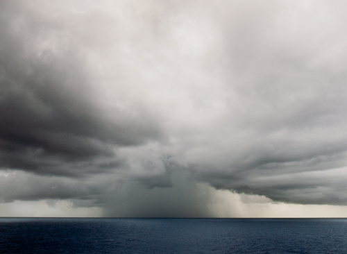 A stark landscape, mostly darkening sky over deep blue water, with a storm at the center horizon line.
