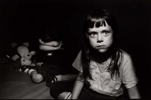 A black and white photograph of two children sitting on a bed. left figure's arms cross face and right figure stares at viewer