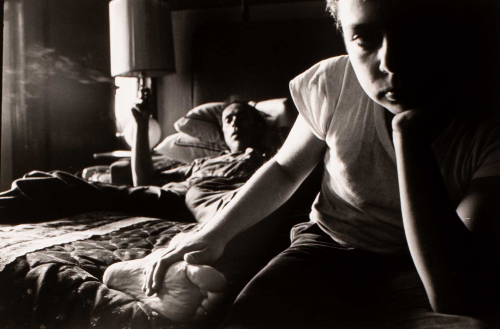 black and white. two male figures on a bed. figure on left lying down and smoking. figure on right is looking at viewer  