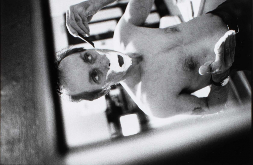  black and white photograph of a shirtless male figure shaving in front of a mirror. The vertical image is horizontally placed