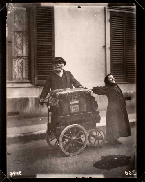Somber bearded man and smiling young girl arms outstretched near cart in the street.  