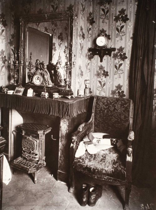 Interior with flowered wall paper, mantle, mirror, and stove on left, chair, boots on left