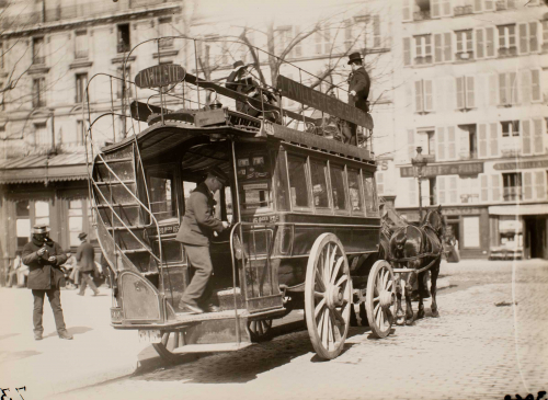 Horse-drawn trolley. Conductor on back of trolley, two passengers on top, buildings in back, pedestrians on left