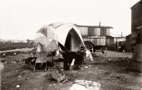 Vagrant's wagon in center surrounded by personal effects.  Other wagons and shacks at right and shacks seen to left on horizon. 