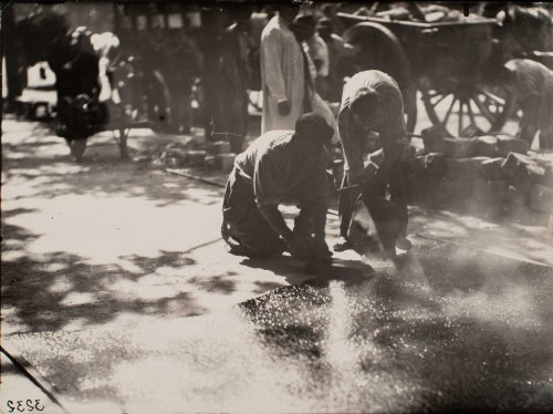 Two men crouched over on a street, finishing concrete, people walking by in background, horse- drawn carts