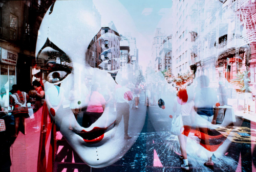 Two large clown masks super-imposed on a street market scene with shoppers in middle of street with buildings lining sides