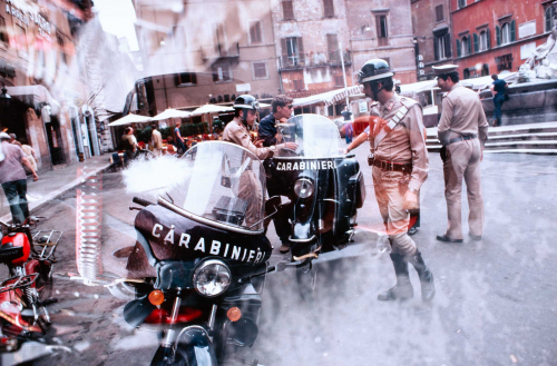 Horse head super-imposed on a scene with two carabinieri and their motorcycles in center with three other figures.