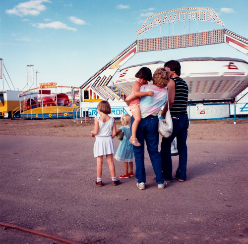  Viewed from behind two young girls in center with woman holding another girl and a man to the right. Carnival rides in back.