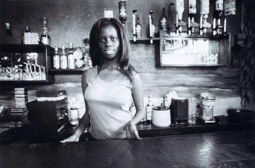 young woman standing behind a counter in a bar with various glasses, liquor dispensers, and compact discs arranged in back