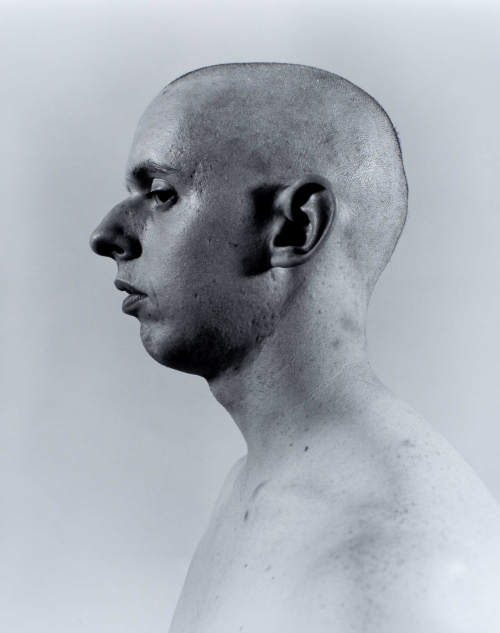 black and white self-portrait bust in profile facing left. The photographer's head is shaved bald and backdrop is light/white.