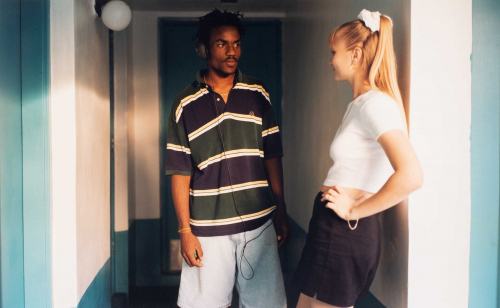 Blue and white hallway with male figure in striped shirt in middle of picture plane. Left-facing blonde female leaning on wall