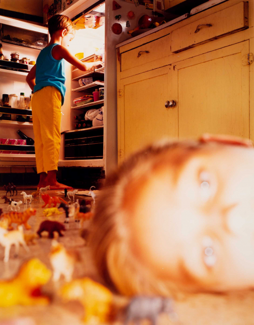 Blurry image of a child's face up close; another child in the background with refrigerator open; several toys sprawled on floor