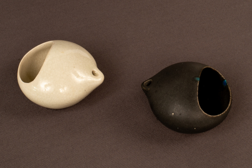 2 small egg-shaped sculptures, one transluscent white and one flat black, each with a tiny spout and an ovoid opening 