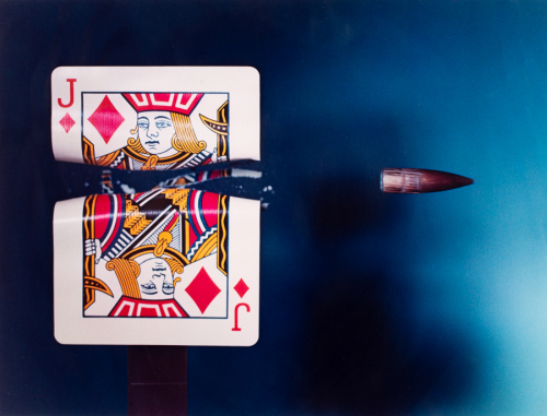 Color photograph of a bullet and a jack of diamonds playing card moments after the bullet has severed the card in two