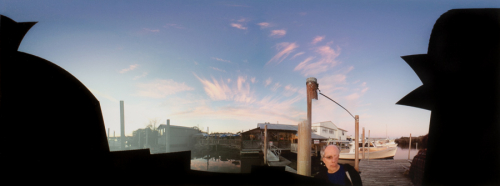 panorama, a sunset at a marina with wispy clouds overhead and a truncated figure in the lower right. 