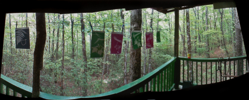 Small red and green prints hang on a line with clothespins from a green-railed porch against a forest backdrop.