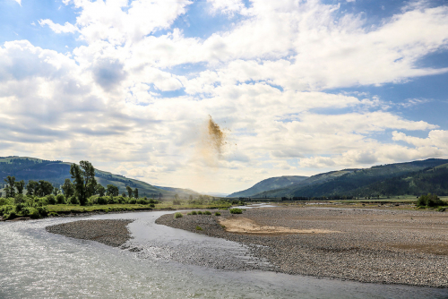 A landscape image of of a pebbly stream and a cloudy blue sky. In the center of the image is a spray of brown dust.