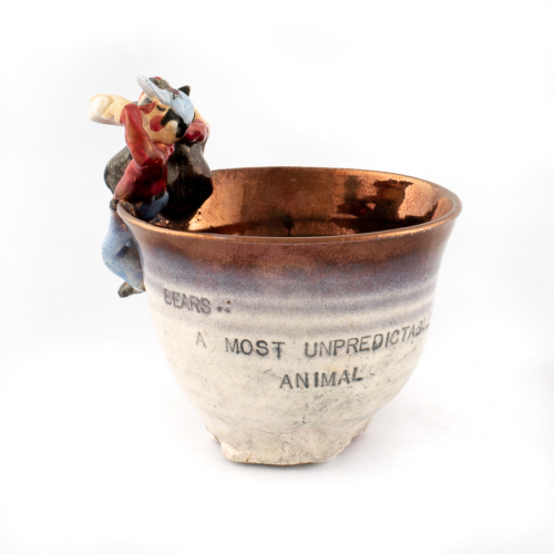 mostly white cup with text and a bronze glazed inside. Bear and a man in jeans, red shirt, and cap (both drinking) on cup's lip.
