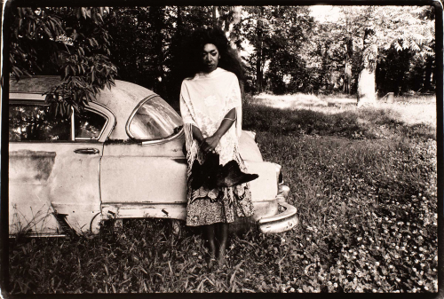 A black and white photograph of a female figure in a wooded landscape holding a dead bird and leaning against an abandoned car.