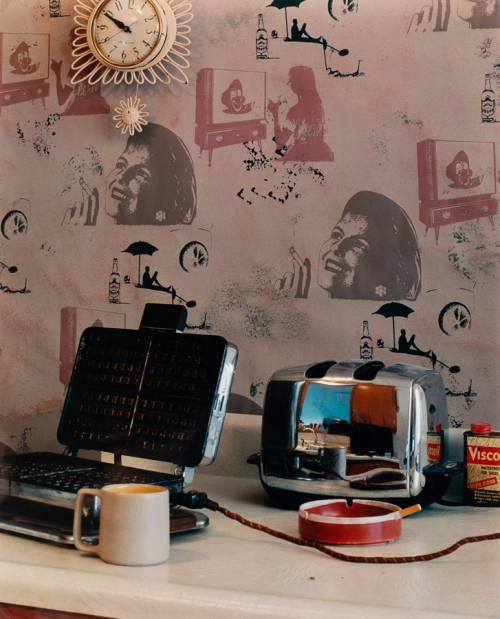 Various kitchen objects, ashtray, and shoe polish on counter. Wallpaper with various printed images, including smoking woman.