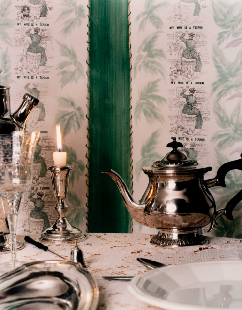  a fancy table setting, with a candle, a teapot and silver; background of wallpaper with "My Wife is a Terror"