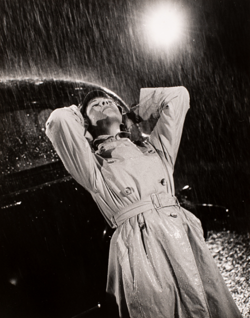 woman wearing trenchcoat with hands behind her neck, leaning to back on a car, it's raining