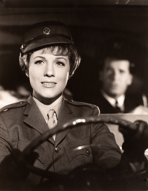 Woman dressed in dark military uniform and hat behind a steering wheel sitting to left of image.  Unfocused image of man behind 