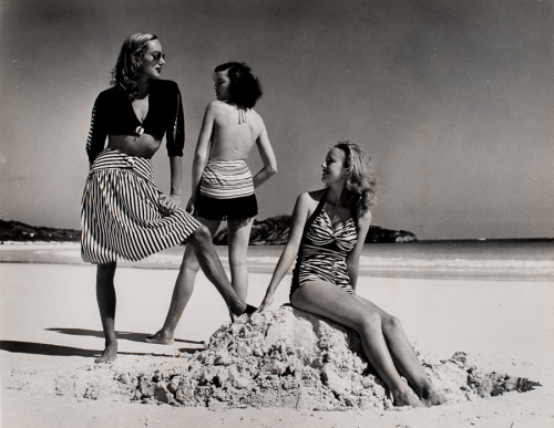 Three women on beach. Left woman stands with her foot on sand pile. middle facing away looking back. right sits on sand pile