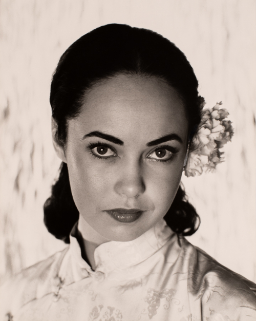 Head shot of woman-not smiling-with dark hair pulled back and flower tucked behind her left ear. 