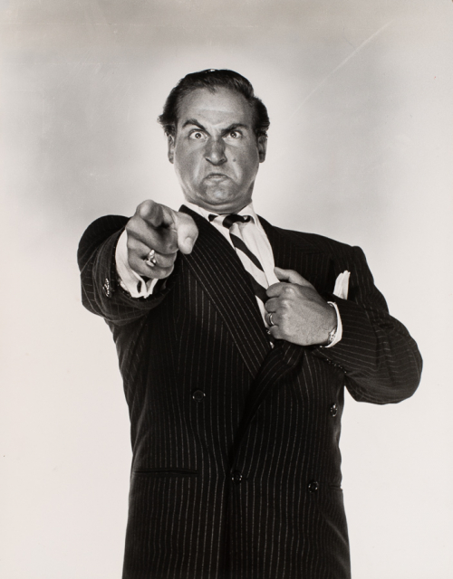 Man in pin-stripe suit, left hand pulling on suit, right hand pointing at viewer, frowning with eyebrow raised and eyes crossed