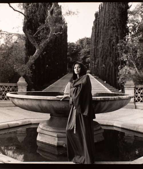 Woman wearing dark cloak with hood up.  Body is turned towards the left, face turned to viewer, leaning on large fountain