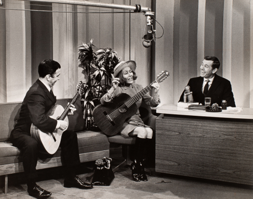Image of people on a talk show set, two people on the left look to be playing guitar and signing, man on right behind a desk