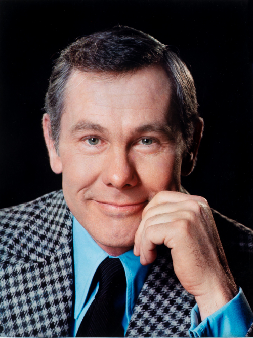 Bust-shot of man with his left hand touching left side of his face smiling. Wearing houndstooth jacket, blue shirt, black tie