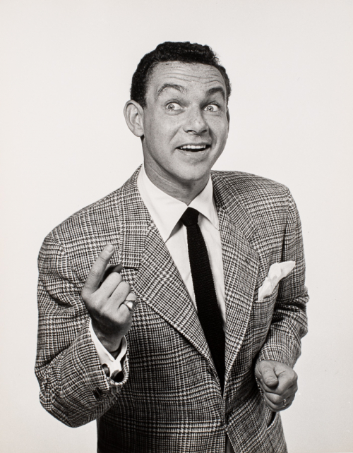 View of man from waist and up wearing plaid jacket and dark tie. Facing slightly to the right, his left hand pointing up