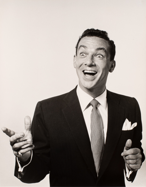 Man in suit with smiling almost surprised looking facial expression holding hands out as if pointing or gesticulating