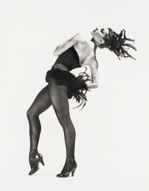 Woman wearing an all black dancing ensemble with a feather bustle and feathers in her hair, torso bending back