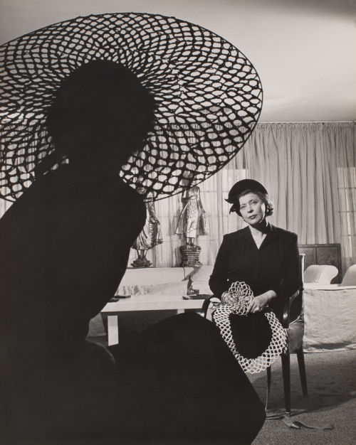 Profile of seated woman in large hat facing right and away from viewer along left side of image. Another seated woman at back 