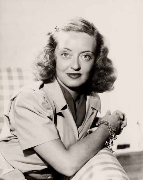 Woman from waist up seated on chair leaning on arm with hands clasped shoulders tilted to right head facing viewer