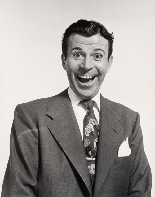Bust-shot of man in medium-dark suit and loud tie with big smile. 