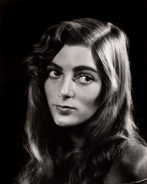 Head-shot of woman with long hair turned towards left side of image with head and eyes turned to the right.