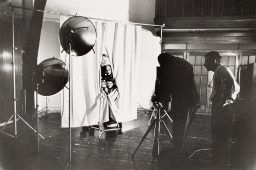 Interior of photography studio. Three people looking out from behind two curtains left of center of image.