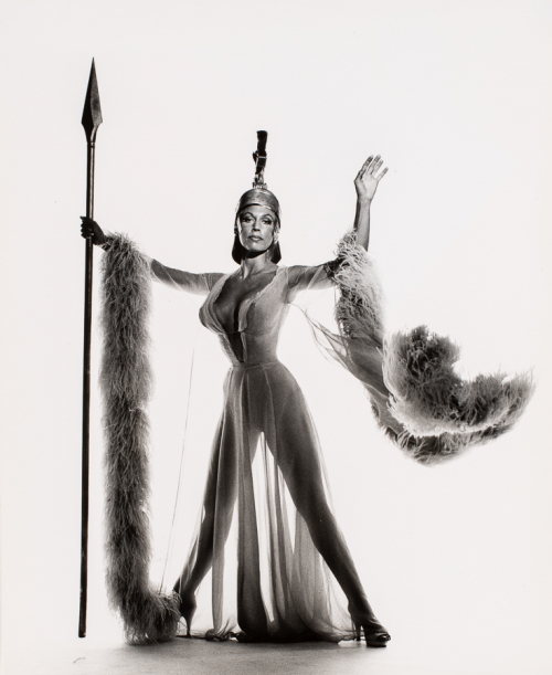 Woman facing viewer in wide stance with arms extended out. Right arm holds a spear. She is wearing helmet and sheer gown 