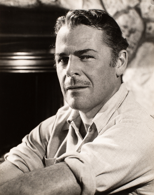Man whose body is positioned facing the left of image with face turned facing viewer. wearing button up with sleeves rolled up