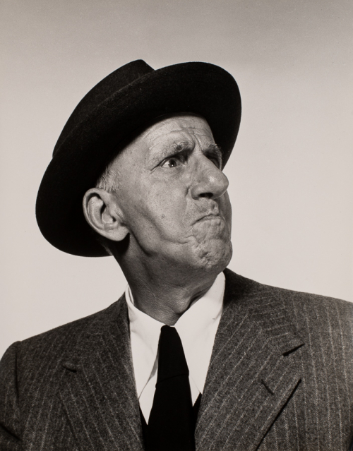 Man with torso facing viewer and head facing the right, his lips are pressed together like angry wearing suit and hat
