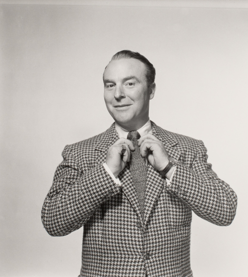 Man from waist and up in a hounds tooth jacket, adjusting his tie. His body is facing the viewer but his head is turned slightly